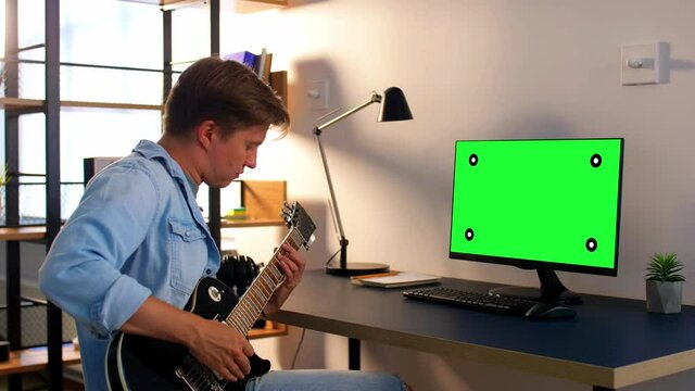 leisure, music and people concept - young man or musician with green chroma key screen on computer playing bass guitar sitting at table at home