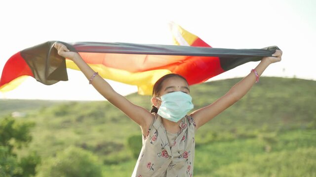 Little girl in medial mask with closed eyes enjoying fresh air by holding German flag in hand on top of mountain.