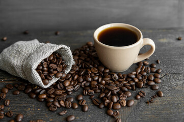 Coffee scattered fron a linen bag and a cup of black coffee Espresso or Americano standing on a wooden table. Fresh arabica coffee beans on linen textile. Aromatic freshly brewed coffee.