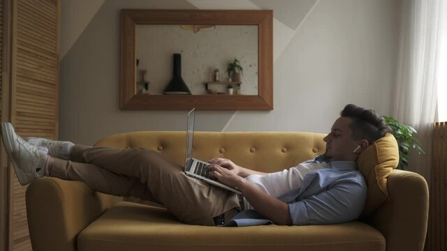 A young man is lying on the sofa with a computer on his lap and working