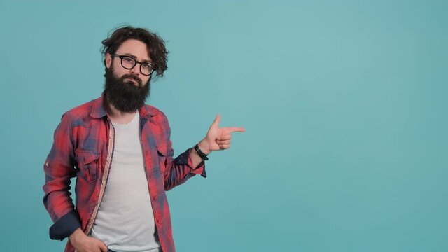 A confident looking young bearded man is pointing to the left and nods, standing isolated over turquoise background.