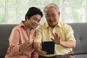 Senior Asian elderly couple in home casual outfit with happy smiling emotion sitting in living room using smart tablet to video call facetime with family