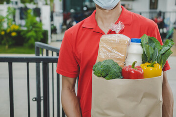 smart delivery man with protection face mask in red uniform holding fresh food set bag waiting for customer at door home, healthy food, express delivery, food delivery, online shopping concept