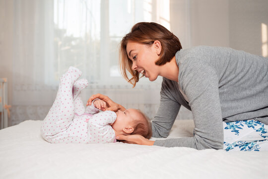 A Young Happy Mother Is Playing With Her Baby While Lying On The Bed. Side View. Concept Of Motherhood And Child Care