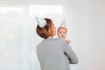 A young mother holds a cute baby in her arms. Festive paper hats on their heads. Rear view. Window on the background. Concept of motherhood and happy birthday of the child