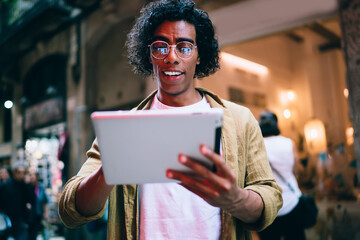 Positive caucasian male with curly hair share content in blog on digital tablet outdoors near neon...