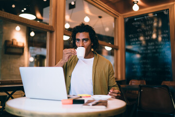 Serious handsome businessman enjoying caffeine beverage for refreshment working remotely in cafeteria, pensive young male having coffee break during distance job on online project via laptop
