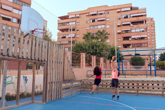 Two young girls with protective face mask playing basketball on a city street court, one throwing and the other trying to block out.
