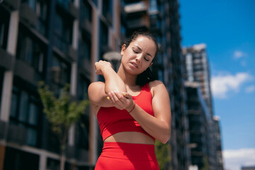 Front view of beautiful young woman in red bra feeling pain in her elbow, hand. during sport workout outdoors. Close-up view. Blurred apartment blocks on background.