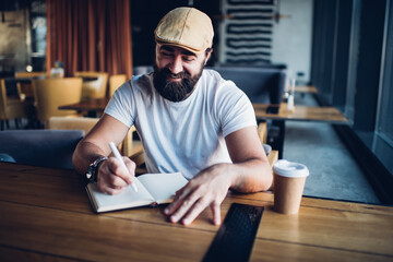 Prosperous bearded mature male author satisfied with work in cafe interior writing in notebook,...