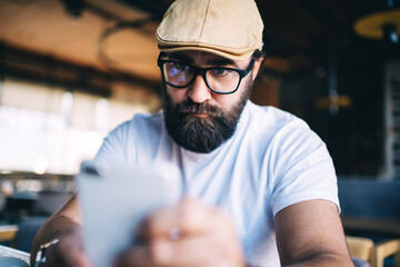 Serious caucasian bearded male in eyewear for vision correction reading news from social networks, blurred smartphone in hand of pensive man 40s blogger checking mail and sending text messages