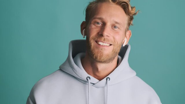 Attractive stylish blond bearded man in hoodie and wireless earphones happily looking in camera over colorful background 