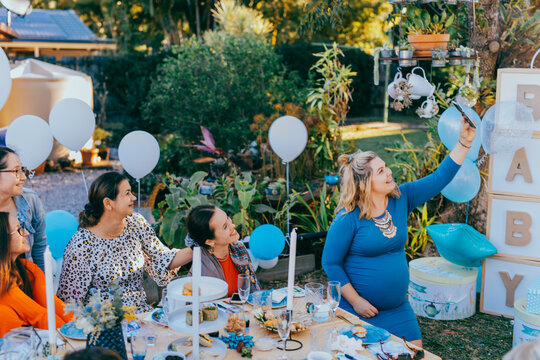 Female friends taking selfie with pregnant woman at a baby shower. Mobile photography, party decorations in white and blue colors, baby boy
