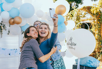 Female friends taking selfie with pregnant woman at a baby shower. Mobile photography, party decorations in white and blue colors, baby boy - obrazy, fototapety, plakaty