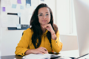 Successful young businesswoman working from home or in office. Woman with long brown hair wear bright yellow blouse and round glasses . Looking at camera. Her face propped on her hand.