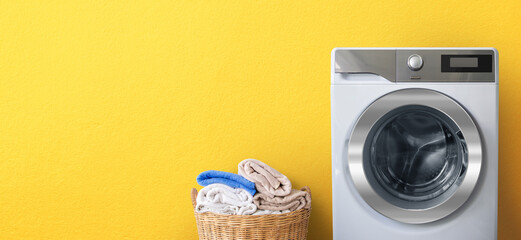Washing machine with laundry near clean bath towels in wicker basket on yellow wall background-Copy...