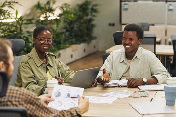 Portrait of two young African-American people smiling at colleagues while sitting at table during meeting in modern office and discussing work project