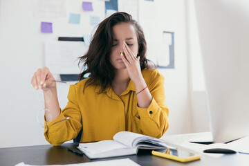 Stressed office young woman holding glasses in her hand. Woman touching her face being tired. Female freelance worker. Notes and plans on background. Exhausted person.