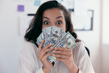 Businesswoman with long brown hair in formal wear white shirt holding lots of dollar banknotes. Happy woman holding money in office. Office job or work at home concept.