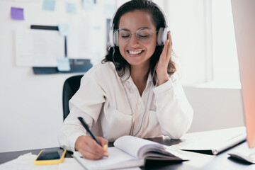 Pretty young caucasian woman in headphones dancing to music and writing lists in her planner. Phone and paper on black table. Positive thinking person concept. Female remote of office worker.
