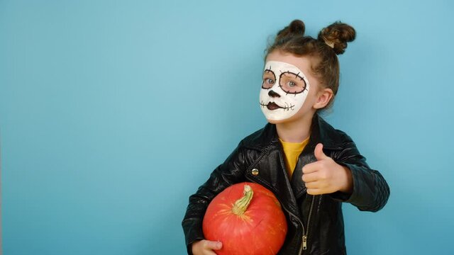 Child girl has face arfully painted to resemble skulls holds orange pumpkin,  shows sign ok, wears black leather jacket, isolated on blue studio background with copy space. Happy Halloween concept