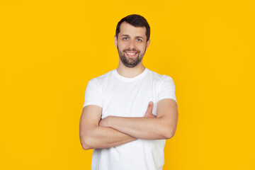 Young man with beard in white t-shirt happy face smiling with crossed arms looking at the camera. Positive man stands on isolated yellow background