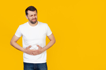 Fototapeta na wymiar A young man with a beard in a white T-shirt with a hand on his stomach because of indigestion, feeling sick. Pain concept. Stands on isolated yellow background