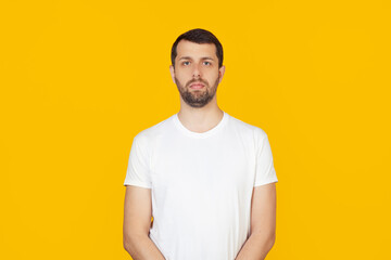 Young man with a beard in a white t-shirt with a serious expression on his face. Simple and natural looking into the camera. Stands on isolated yellow background