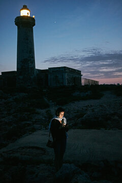 jeune femme regardant son smartphone dans la nuit en bord de mer à côté d'un phare en Sicile