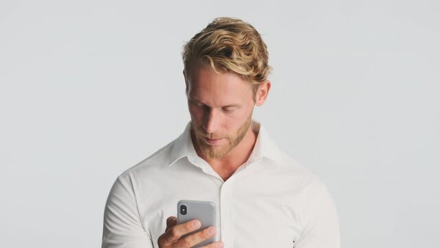 Attractive serious blond bearded man in shirt intently using smartphone waving no gesture on camera over white background