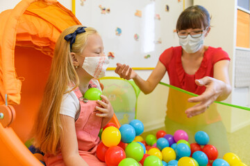 Toddler girl in child occupational therapy session doing playful exercises with her therapist during Covid - 19 pandemic, both wearing protective face masks.