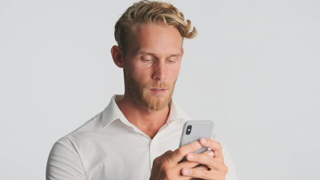 Handsome blond bearded businessman in shirt intently working on smartphone and smiling over white background