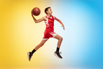 Fototapeta na wymiar Jump high. Full length portrait of young basketball player in uniform on gradient studio background. Teenager confident posing with ball. Concept of sport, movement, healthy lifestyle, ad, action