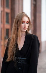 A girl with red hair, dressed in a black jacket, posing against the backdrop of a white building. Looking away.