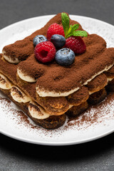 portion of Classic tiramisu dessert with raspberries and blueberries on dark concrete background
