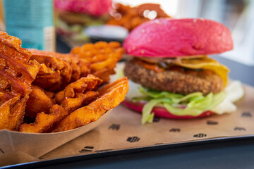 Selective focus on sweet potato waffle cut fries in front of pink beetroot gluten-free hamburger bun with vegan black beans patty, nachos, jalapeño, lettuce and Mexican sauce