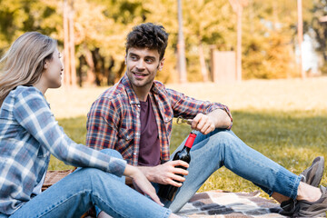 man holding bottle of red wine near woman sitting on plaid blanket in park