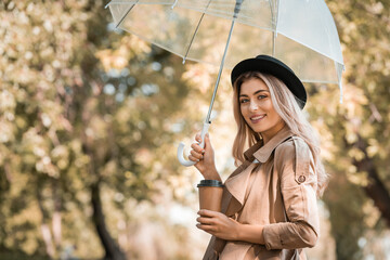 blonde woman in hat holding umbrella and disposable cup with coffee to go in autumnal park