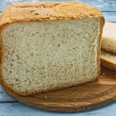 Sliced loaf of white bread on a wooden cutting board. Nearby are two slices. Crispy golden crust and porous crumb. Background - blue painted boards of the table.
