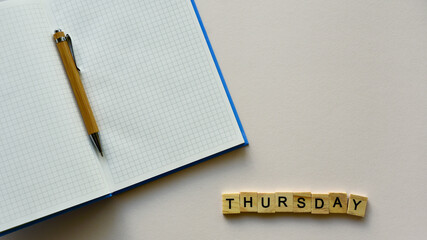 Notepad with pen and wooden word "Thursday" on a beige paper background. Top view. Close-up. Flat lay.