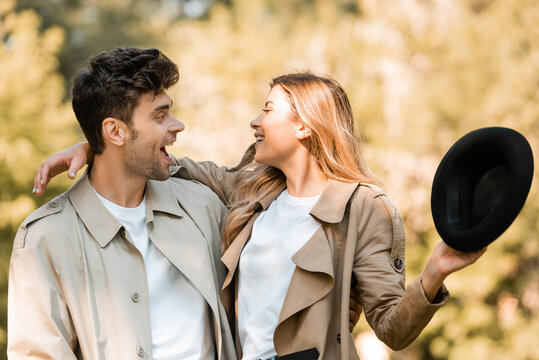 excited woman holding hat and looking at boyfriend in autumnal park