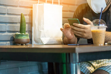 Asian male customer wear face mask using smartphone while sit in waiting social distance queue for take away drink in coffee shop. New normal lifestyle concept. Selective focus on smartphone