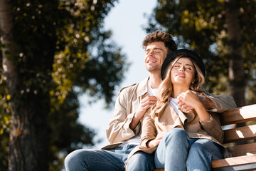 Naklejka premium blonde woman in hat and man in trench coat sitting on wooden bench and holding hands