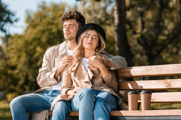Naklejka premium blonde woman in hat and man in trench coat sitting on bench near paper cups