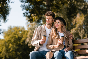 Fototapeta premium blonde woman in hat and man in trench coat holding disposable cups while sitting on bench
