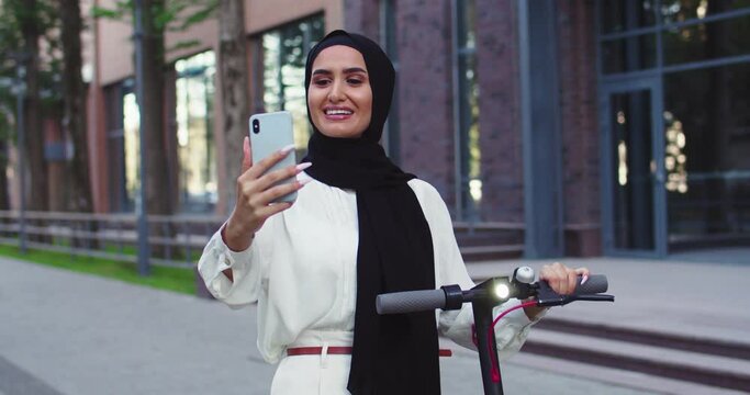 Young Arabic beautiful women in traditional headscarves, influencer shooting video blog, making selfie with electric scooter, meeting a friend on the street. Pretty Arabian females in hijabs at city.