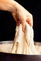 Hand stretches bread dough from a bowl on dark background