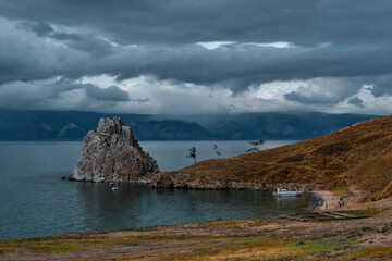 Cape Shamanka rock in blue Lake Baikal among the grassy steppes, against the background of mountains and dark clouds