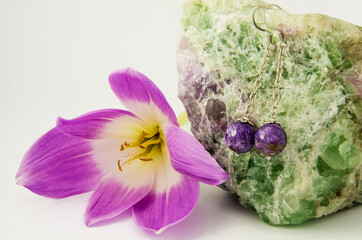 Pendant earrings on a chain with charoite beads hang on a green fluorite near a pink flower.