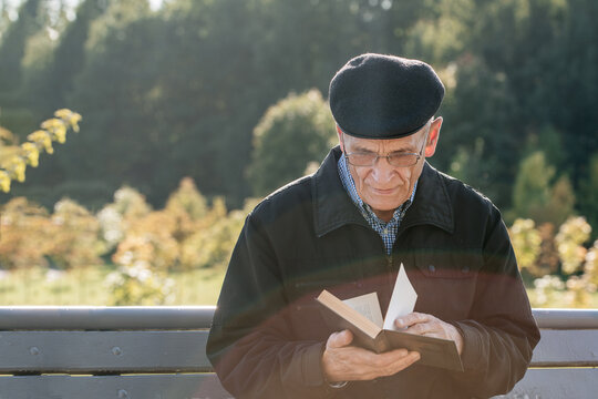 Elderly Mature Man Wearing Glasses And Black Cap Sitting On Wooden Bench And Reading Old Book